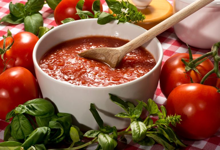 Bowl of fresh healthy homemade tomato puree with a wooden spoon for use in Italian cuisine surrounded by fresh basil for seasoning and ripe red tomatoes